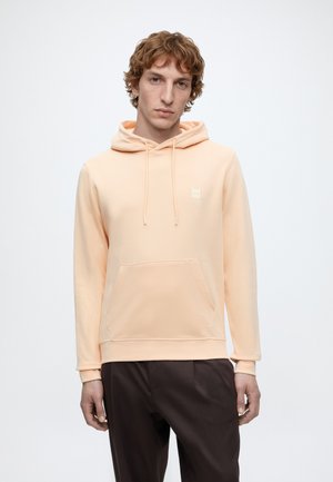 Junger Mann trägt einen hellpfirsichfarbenen Hoodie mit Fronttasche und Kordelzug, kombiniert mit dunklen Hosen, steht vor einem schlichten weißen Hintergrund.