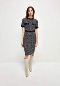 Vestido de punto en tweed blanco y negro con mangas cortas, que presenta dos bolsillos frontales y una silueta ajustada, combinado con botas de rodilla beige.