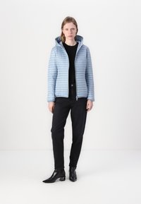 Lichtblauwe pufferjack met capuchon, voorzien van een geweven textuur en een voorkant met rits. Zwarte broek en enkellaarsjes met hak maken de outfit compleet.