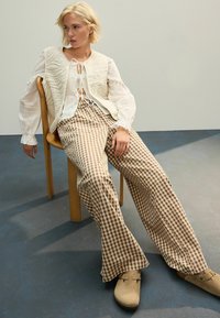 Beige vadderad väst över en vit blus med knytband, kombinerad med bruna rutiga utsvängda byxor och beige träskor, sittande på en trästol.