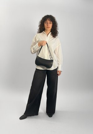 Sac à main en cuir synthétique noir avec une forme incurvée, garnitures argentées, tenu par une personne portant une chemise blanche boutonnée et un pantalon noir à larges jambes.