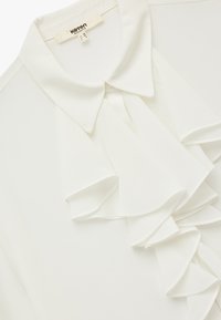 Hvid bluse med krave og kaskader af flæser ned ad fronten. Fremstillet af let materiale, med en glat overflade og knaplukning.