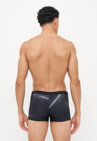 Schwarze Badeshorts mit gepunktetem Muster, elastischem Bund und figurbetontem Design. Das Material wirkt glatt mit einem leichten Glanz.