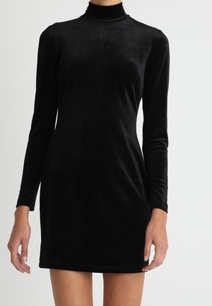 Robe courte en velours noir à manches longues avec un col montant portée par une personne debout devant un fond clair uni.