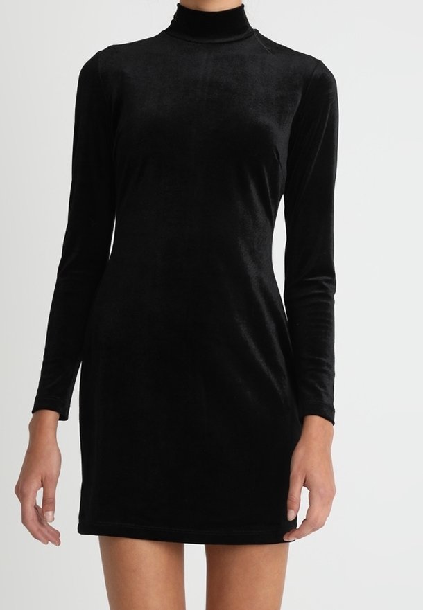 Robe courte en velours noir à manches longues avec un col montant portée par une personne debout devant un fond clair uni.