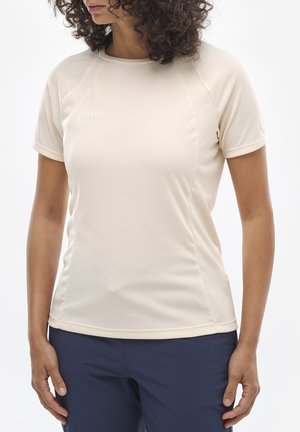Femme aux cheveux bouclés portant un t-shirt beige à manches courtes et col rond, et un pantalon bleu marine, debout avec les bras détendus le long du corps.