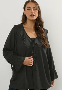 Svart långärmad blus med volangkrage, knytfram och elastiska ärmar. Mjukt tyg med en flödande silhuett.