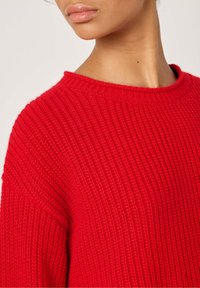 Pull en tricot rouge avec une texture côtelée, col rond et épaules tombantes. Affiche un design épais et confortable, idéal pour le superposition.