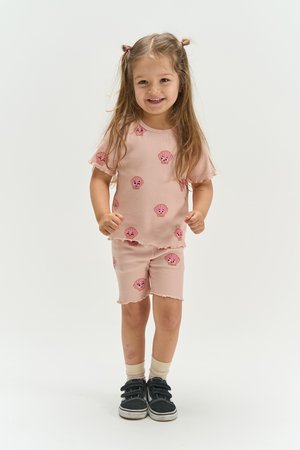 Smiling young girl with long hair in pink matching shorts and shirt with expressive face prints, wearing black sneakers on a white background.