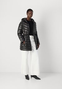 Doudoune noire avec capuche, design matelassé, taille élastique et poches latérales, assortie à un pantalon large blanc et des talons noirs.