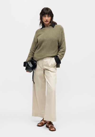 Femme portant un pull en maille vert olive par-dessus une chemise noire, pantalon large crème, sandales marron, tenant un sac noir, debout sur fond blanc.