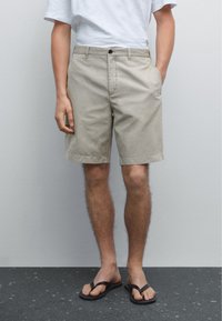 Beige Baumwollshorts mit lockerer Passform, zwei Seitentaschen und Knopfverschluss. Getragen mit einem weißen T-Shirt und schwarzen Flip-Flops.