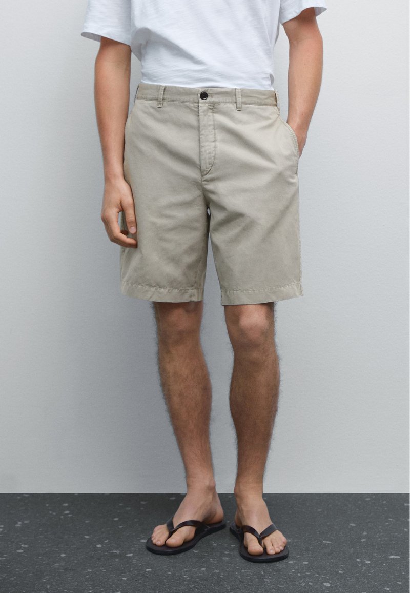 Beige Baumwollshorts mit lockerer Passform, zwei Seitentaschen und Knopfverschluss. Getragen mit einem weißen T-Shirt und schwarzen Flip-Flops.