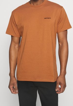 Mann trägt ein schlichtes braunes Kurzarm-T-Shirt mit kleinem schwarzem Carhartt-Logo auf der linken Brust und schwarze Hose.