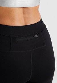 Leggings noirs taille haute en matériau extensible avec une texture lisse. Présente une petite poche zippée à l'arrière.