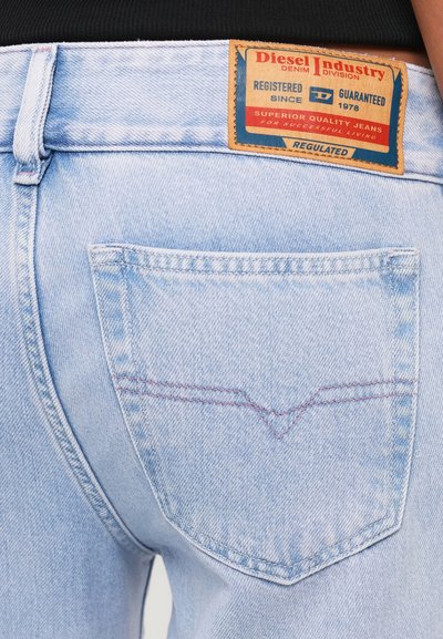 Jean en denim bleu clair avec une poche arrière bien visible, des détails de couture rouges et une étiquette beige contenant des informations sur la marque au niveau de la taille.