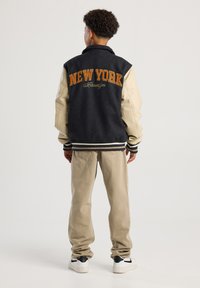 Person wearing a black and beige varsity jacket with "New York Brooklyn" on the back, beige pants, and white sneakers, standing facing away.