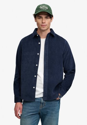 Man draagt een groene pet, donkerblauw corduroy overhemd met knopen over een wit T-shirt, blauwe jeans en een polshorloge, staand tegen een effen achtergrond.