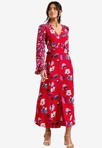 Vestido maxi rojo con flores, con mangas largas de patrón, escote en V y abertura lateral. Presenta diseños de flores moradas y blancas sobre un fondo brillante.