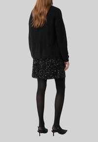 Schwarzer Strickpulli mit lockerer Passform, kombiniert mit einem kurzen schwarzen Paillettenrock. Das Outfit wird durch transparente schwarze Strumpfhosen und hohe Schuhe ergänzt.