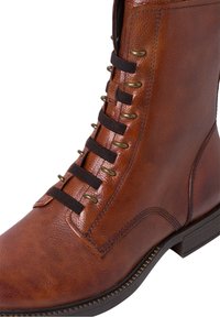 Stivale alla caviglia in pelle marrone con finitura testurizzata, dotato di lacci neri, occhielli in ottone e una suola piatta resistente. Cuciture dettagliate visibili.