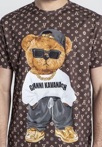 Bruine graphic t-shirt met een beer die een zonnebril, een wit shirt, een gouden ketting en grijze joggers draagt, tegen een patroon achtergrond.