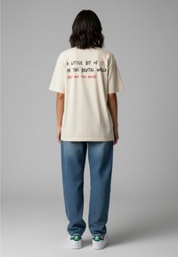 Cremfarbendes Oversized-T-Shirt mit schwarzem und rotem Textdesign auf dem Rücken. Kombiniert mit locker sitzenden blauen Jeans und weißen Sneakers.