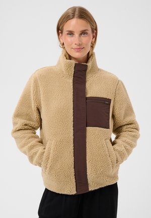 Kvinde iført en beige fleecejakke med en brun lodret stribe og en lynlås brystlomme, hænderne i sidelommerne, vendt mod kameraet med en neutral baggrund.