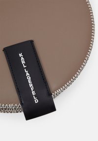 Runde, taupefarbene Kunstledertasche mit silbernem Reißverschluss und schwarzem Lederanhänger, geprägt mit "KARL LAGERFELD" in Weiß.