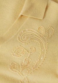Pull en tricot jaune avec un col en V et une broderie décorative ornée de sequins formant un motif tourbillonnant sur le devant.