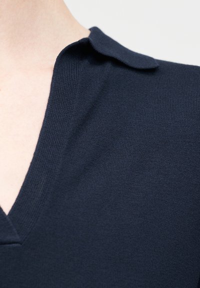Gros plan sur le col d'un vêtement en maille bleu marine sur l'épaule et le cou d'une personne, avec un détail de pli court et souple.