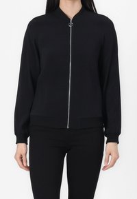 Schwarze Bomberjacke mit Stehkragen, vorderem Reißverschluss und langen Ärmeln mit gerippten Bündchen. Glattes Gewebe mit Seitentaschen.