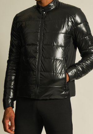 Zwarte glanzende gewatteerde jas met ritssluiting aan de voorkant en ritszakken aan de zijkant, gedragen door een persoon met één hand in de zak.