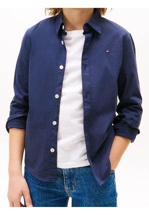 Camisa azul marino abotonada, tela ligera, cuello puntiagudo, mangas remangadas, botones blancos como detalles, combinada con una camiseta blanca y jeans azules.