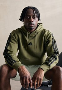 Hoodie verde-oliva com cordão, riscas pretas nas mangas e um pequeno logo preto no peito. Tecido suave, corte descontraído.