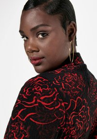 Černý oděv s červeným květinovým vzorem, s texturovaným povrchem. Zlaté kruhové náušnice a hladký krátký účes dotvářejí vzhled.