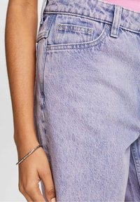 Vaqueros de mezclilla de color púrpura claro con una textura sutil. Presentan bolsillos frontales y un diseño de pierna recta, complementados con una pulsera plateada.