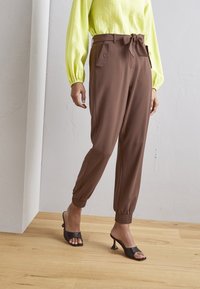 Person i bruna högmidjade byxor med sidofickor och knytbälte, kombinerat med svarta klacksandaler och en gul långärmad topp.