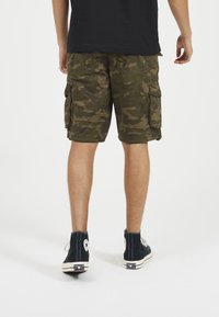 Persona che indossa pantaloni cargo mimetici verdi, maglietta nera e sneakers alte nere Converse, vista da dietro su uno sfondo bianco.