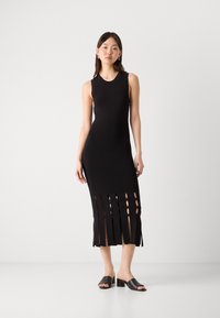 Melna bez piedurknēm kleita, izgatavota no stiepjama auduma, ar vertikālām izgriezumiem apakšmalā. Apvienota ar melnām mulēm ar blokveida papēžiem.