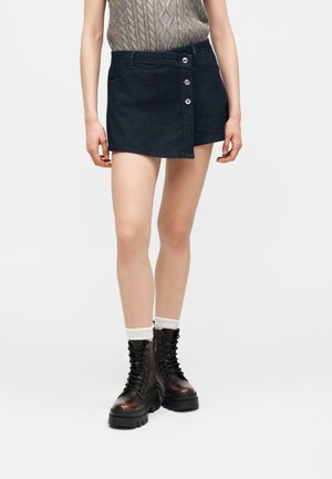 Vrouw draagt een donkere denim minirok met knopen aan de voorkant, een beige kabelgebreide mouwloze top, witte sokken en zwarte chunky veterlaarzen.