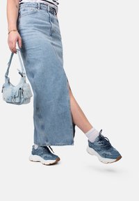 Gonna lunga in denim blu chiaro con uno spacco laterale, abbinata a sneakers blu a fantasia e una borsa a tracolla blu chiaro. Visibili calzini bianchi alla caviglia.