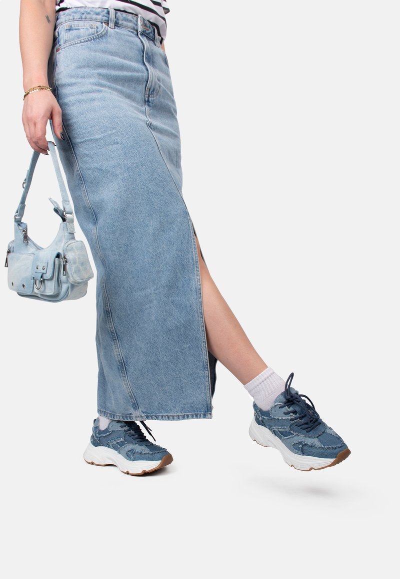 Gonna lunga in denim blu chiaro con uno spacco laterale, abbinata a sneakers blu a fantasia e una borsa a tracolla blu chiaro. Visibili calzini bianchi alla caviglia.