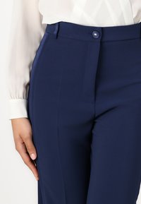 Tamsiai mėlynos pritaikytos kelnės, pagamintos iš lygaus audinio, su plokščiu priekiu, siūlų dekoravimu ir sagčių uždarymu juosmens srityje.