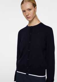 Laivastonsininen neulejakku, jossa on pyöreä pääntie, edessä napitus ja teksturoitu neulos. Sisältää valkoiset raidat helmassa.