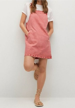 Kobieta w różowej sukience bez rękawów na białym t-shircie, stojąca z rękami w kieszeniach i jedną zgiętą nogą, nosząca beżowe sandały na sznurowadła.