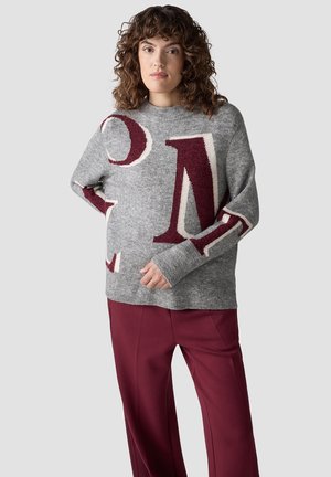 Frau mit lockigem Haar, die einen grauen Pullover mit großen bordeauxroten Buchstaben und passende bordeauxrote Hosen trägt, steht vor einem schlichten Hintergrund.