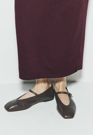 Hnedé kožené Mary Jane baleríny nosené s dlhou tmavou bordovou sukňou, ktoré ukazujú členky a pozadie podlahy.