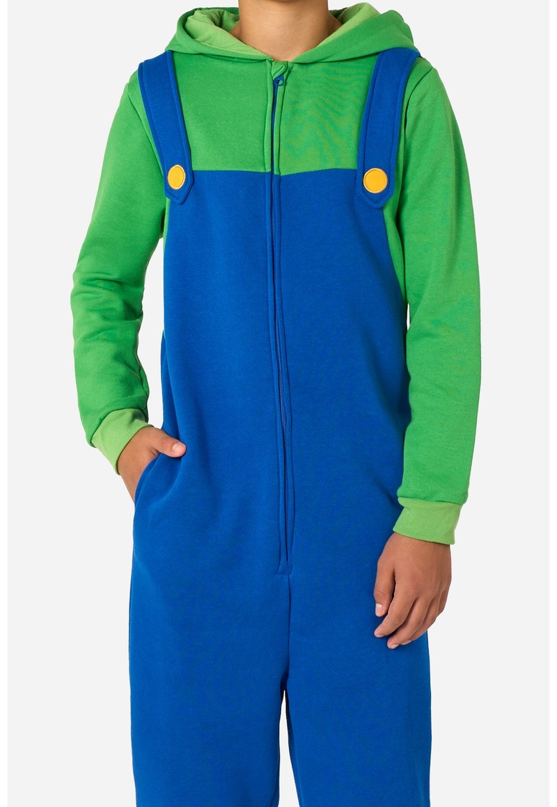 Enfant portant un sweat à capuche vert et une salopette bleue avec des boutons jaunes, les mains reposant à côté et dans la poche, sur un fond uni.