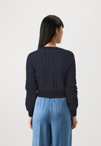 Tmavě modrý svetr s pleteným vzorem a ribbed výstřihem a manžetami, s krátkým střihem a texturovaným vzorem, v kombinaci s světlými džíny.